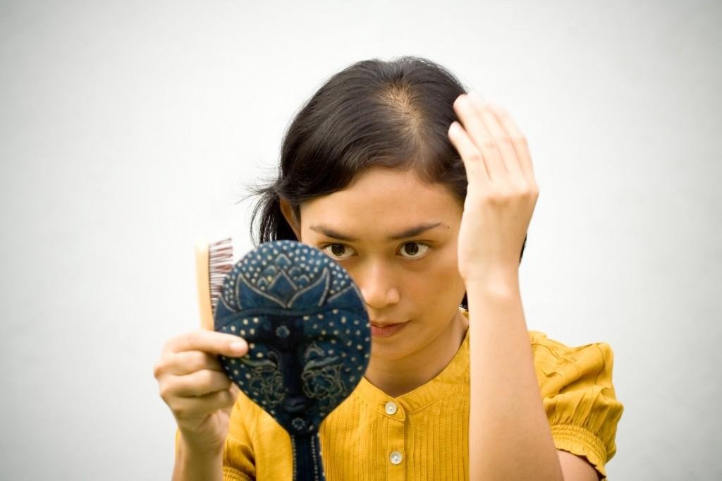 A woman with hair loss looking into a hand mirror, holding a hairbrush and lifting her hair to check her part and scalp