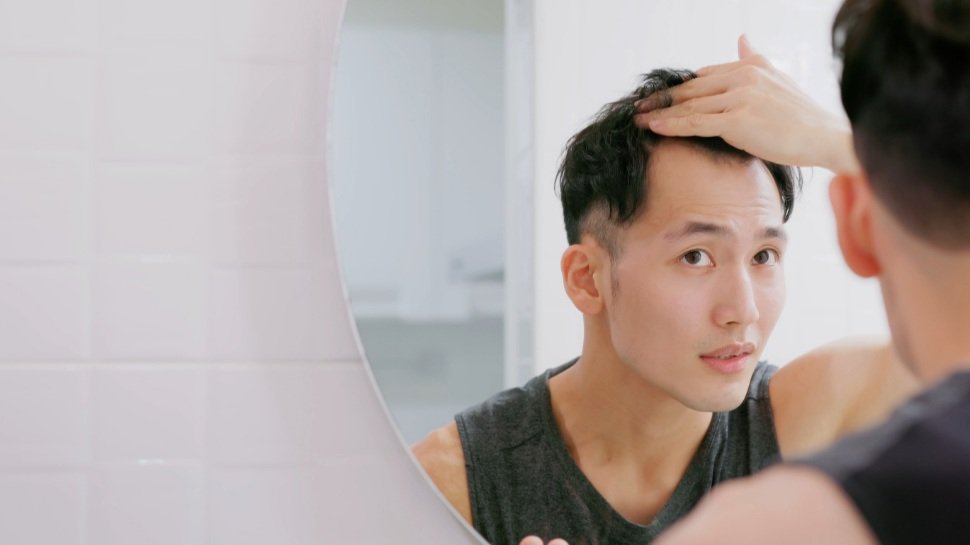 A young man checking his hair for signs of hair loss in a round bathroom mirror, lifting the hair at the front of his head
